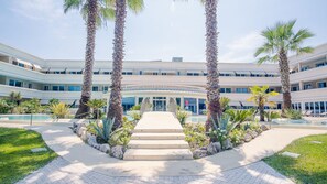 Ein mehrstöckiges Hotel mit Poolbereich in der Mitte, Palmen und einer Treppe zum Eingang.