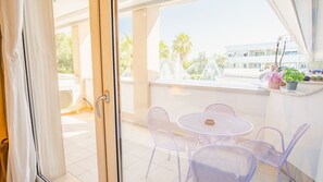 Eine offene Schiebetür zu einem Balkon mit einem weißen Tisch und Stühlen, einem kleinen Topfpflanzen und Blick auf ein Gebäude und Palmen.