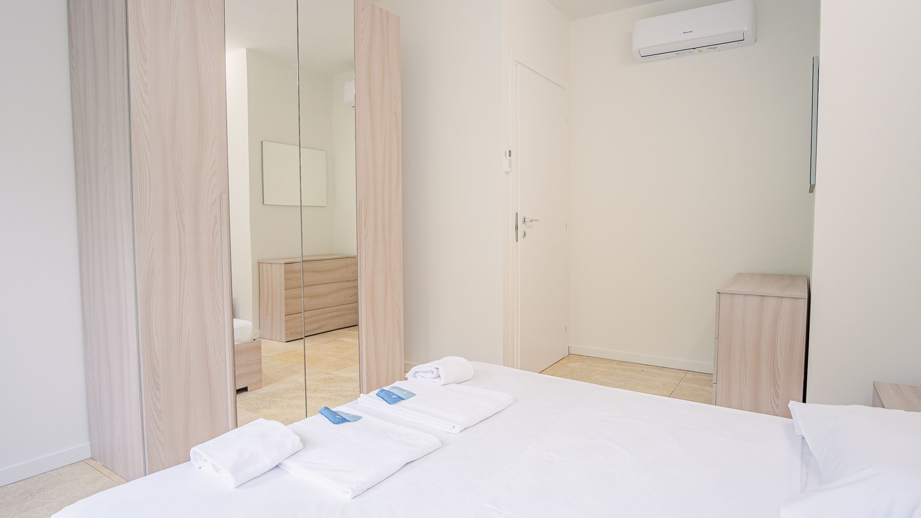 A modern hotel room with a large bed, a wooden wardrobe, a small bedside table, and a mirror.