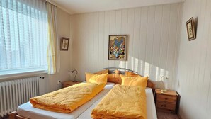 Ein Doppelbett mit gelber Bettwäsche, ein Nachttisch mit Lampe, ein Heizkörper und ein Fenster mit durchsichtigen Vorhängen.