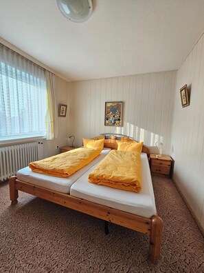 Ein Doppelbett mit gelber Bettwäsche, ein Nachttisch mit Lampe, ein Heizkörper und ein Fenster mit durchsichtigen Vorhängen.