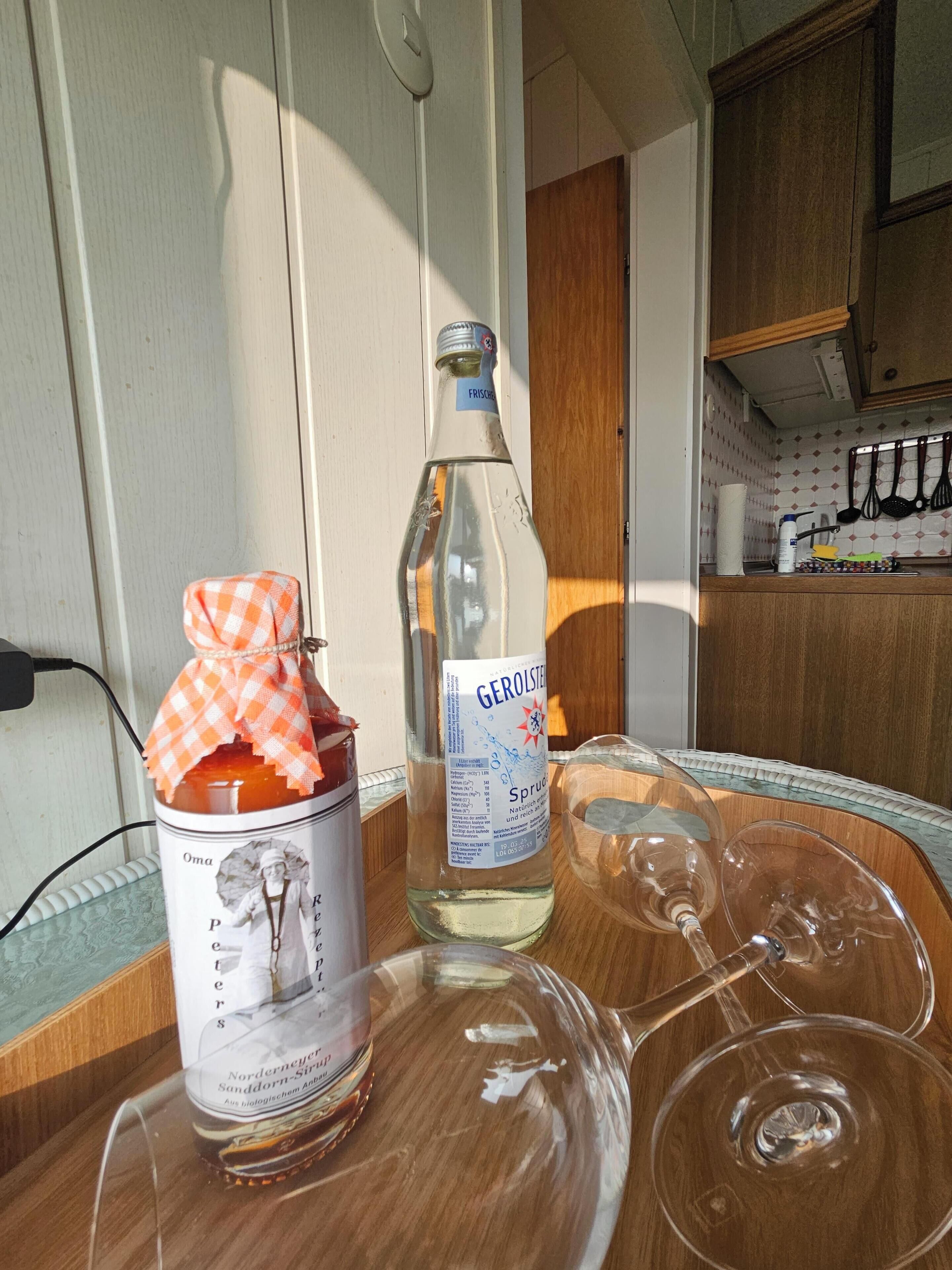 A kitchen area with a bottle of Gerolsteiner water, a jar of jam, and empty wine glasses on a table.