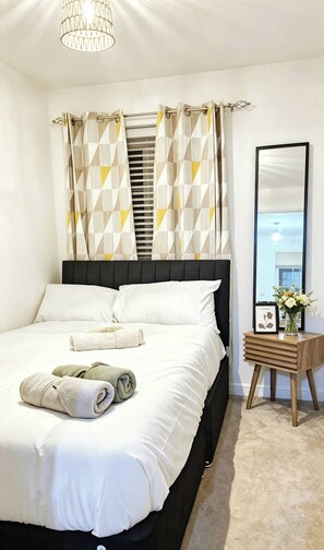 Un dormitorio con cama, almohadas, una mesita de noche con un florero y un espejo.