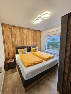 A modern bedroom with a wooden accent wall, a large bed with yellow bedding, and a window with a view of greenery. - Apartment 4 in the Flechtner Building (Norderney)