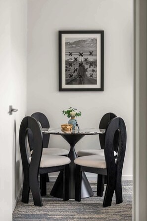 Een eethoek met een ronde tafel, vier stoelen, een vaas met bloemen en een ingelijste foto aan de muur.