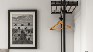 Ein Hotelzimmer mit einem gerahmten Bild an der Wand, einem Nachttisch mit Lampe, einem Kleiderhaken und einer kleinen weißen Bank.