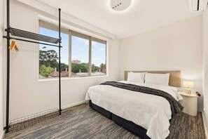 A hotel room with a large bed, a window with a view, a bedside table with a lamp, and a wall-mounted drying rack. - Family Suite with Two King Beds & St Kilda Charm (St Kilda)
