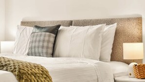 Una cama bien tendida con un edredón blanco y varios almohadones, una lámpara de noche y una manta tejida.