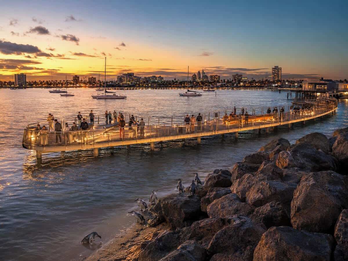 A waterfront promenade with a long, illuminated walkway, boats on the water, and a city skyline in the background.