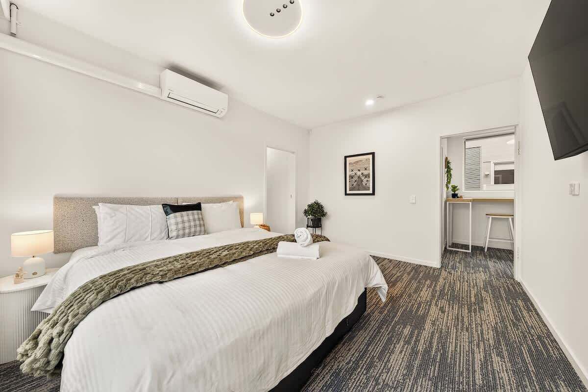 A hotel room with a bed, bedside table, lamp, air conditioning unit, and a television on the wall.