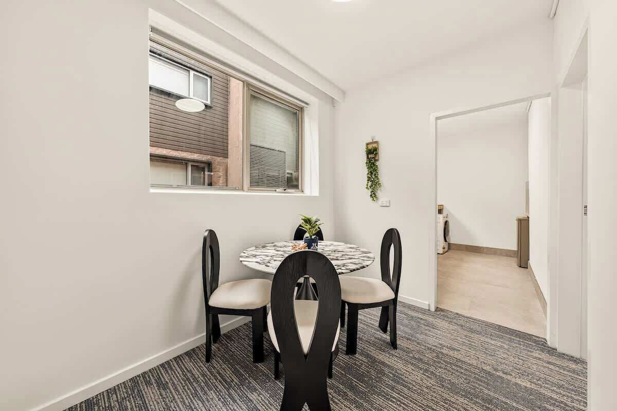 Een eetgedeelte met een ronde tafel, vier stoelen, een raam met jaloezieën en een deur naar een andere kamer.