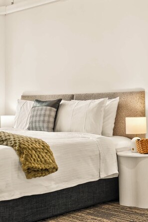 Ein ordentlich bezogenes Bett mit weißer Decke und mehreren Kissen, einer Nachttischlampe und einer gewebten Tagesdecke.