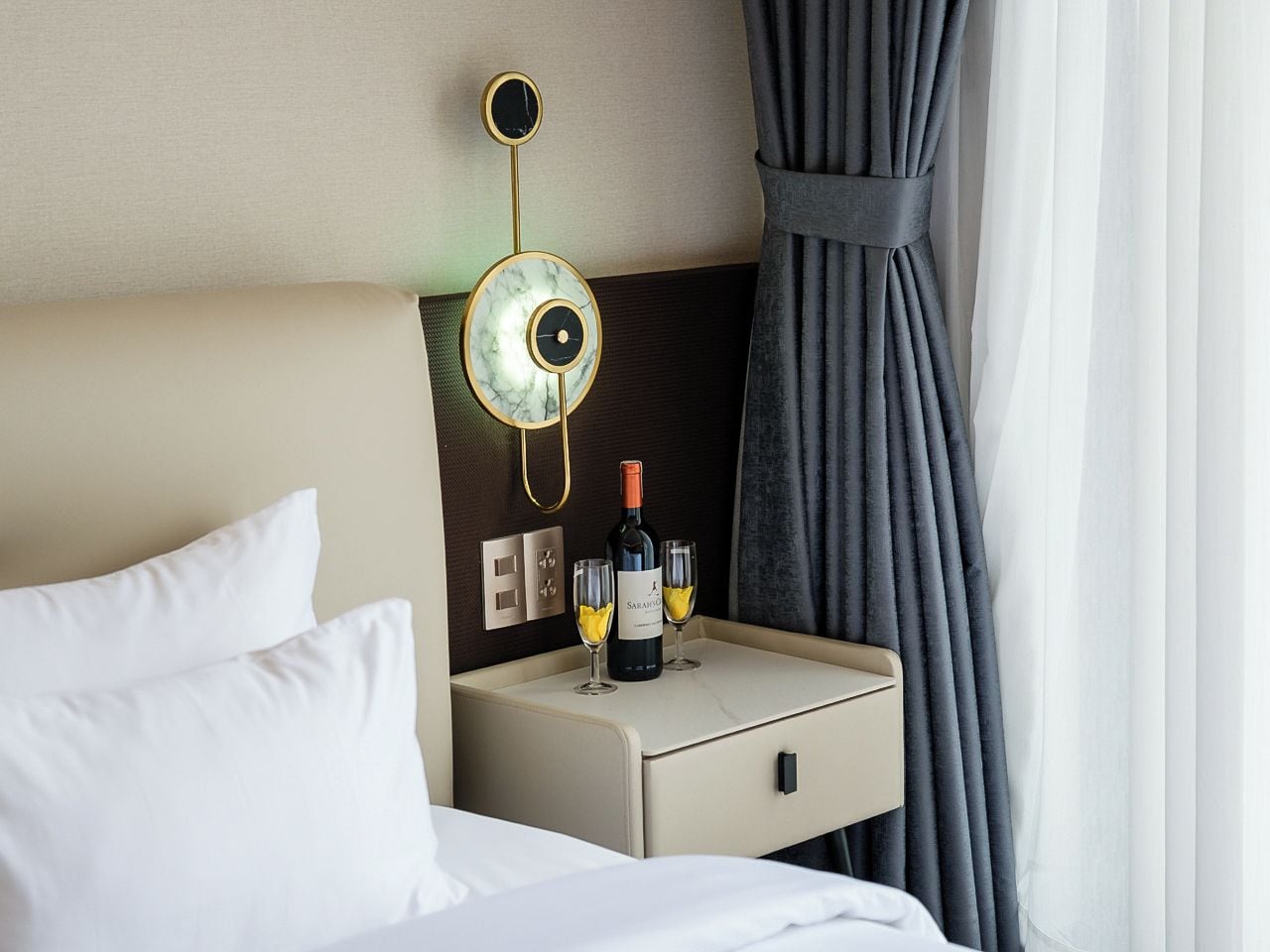 A hotel room with a bed, a nightstand with a bottle of wine and two glasses, a wall clock, and a curtain.