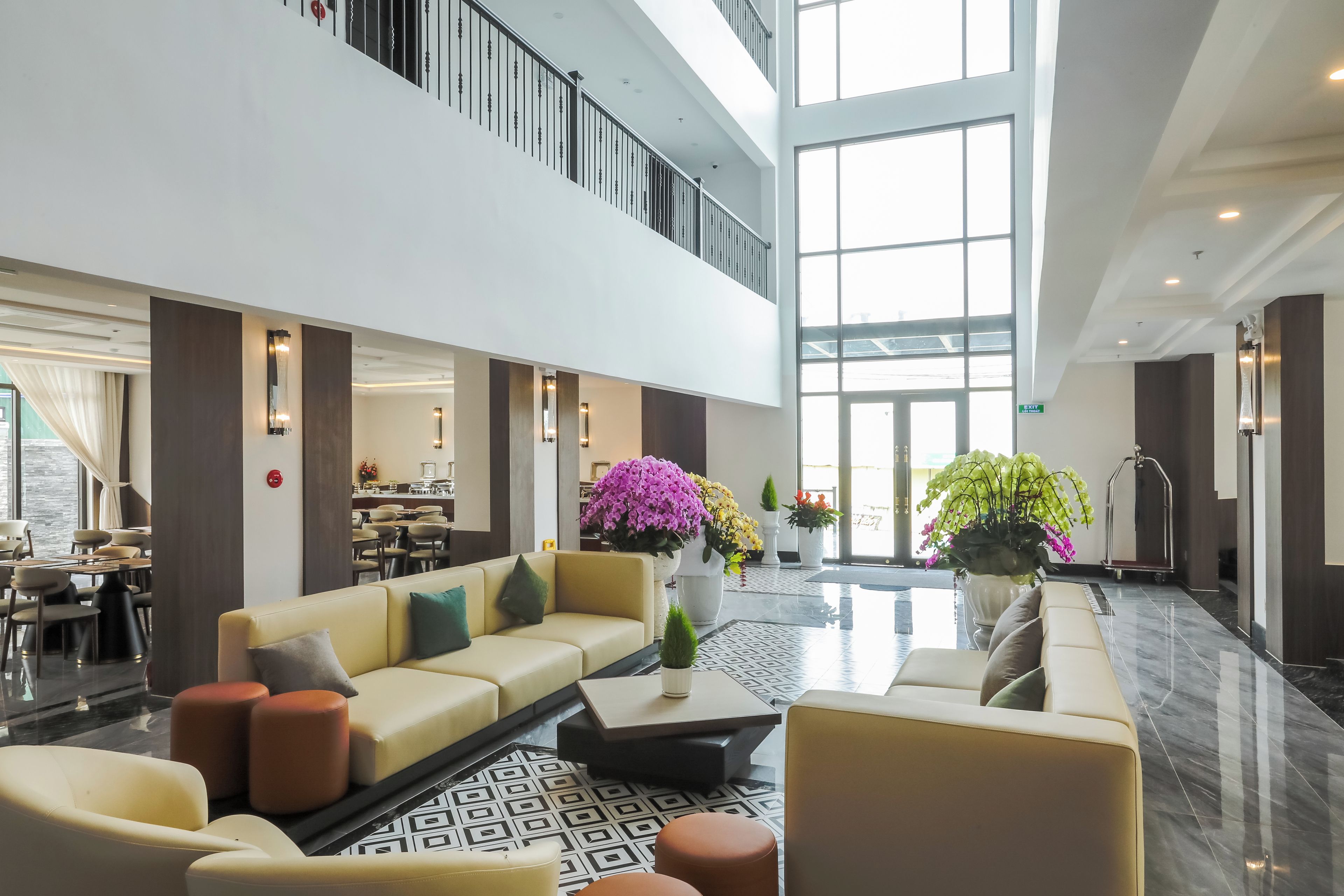 A spacious lobby with a high ceiling, a seating area with sofas and chairs, a dining area in the background, and large windows allowing natural light.
