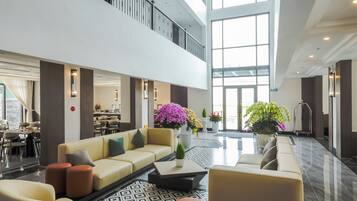 A spacious lobby with a high ceiling, a seating area with sofas and chairs, a dining area in the background, and large windows allowing natural light.