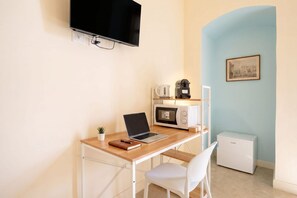 Een compacte kamer met een bureau, een laptop, een koffiezetapparaat, een magnetron, een koelkast, een televisie en een ingelijst kunstwerk aan de muur.