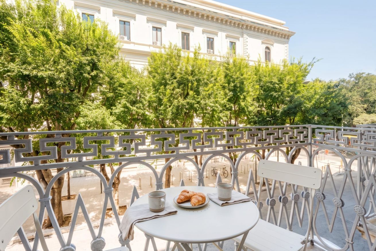 Une table ronde, sur laquelle sont disposés une assiette de croissants et deux tasses, est placée devant une rambarde blanche ornée d’un motif géométrique, d’où l’on aperçoit une rue et un immeuble.
