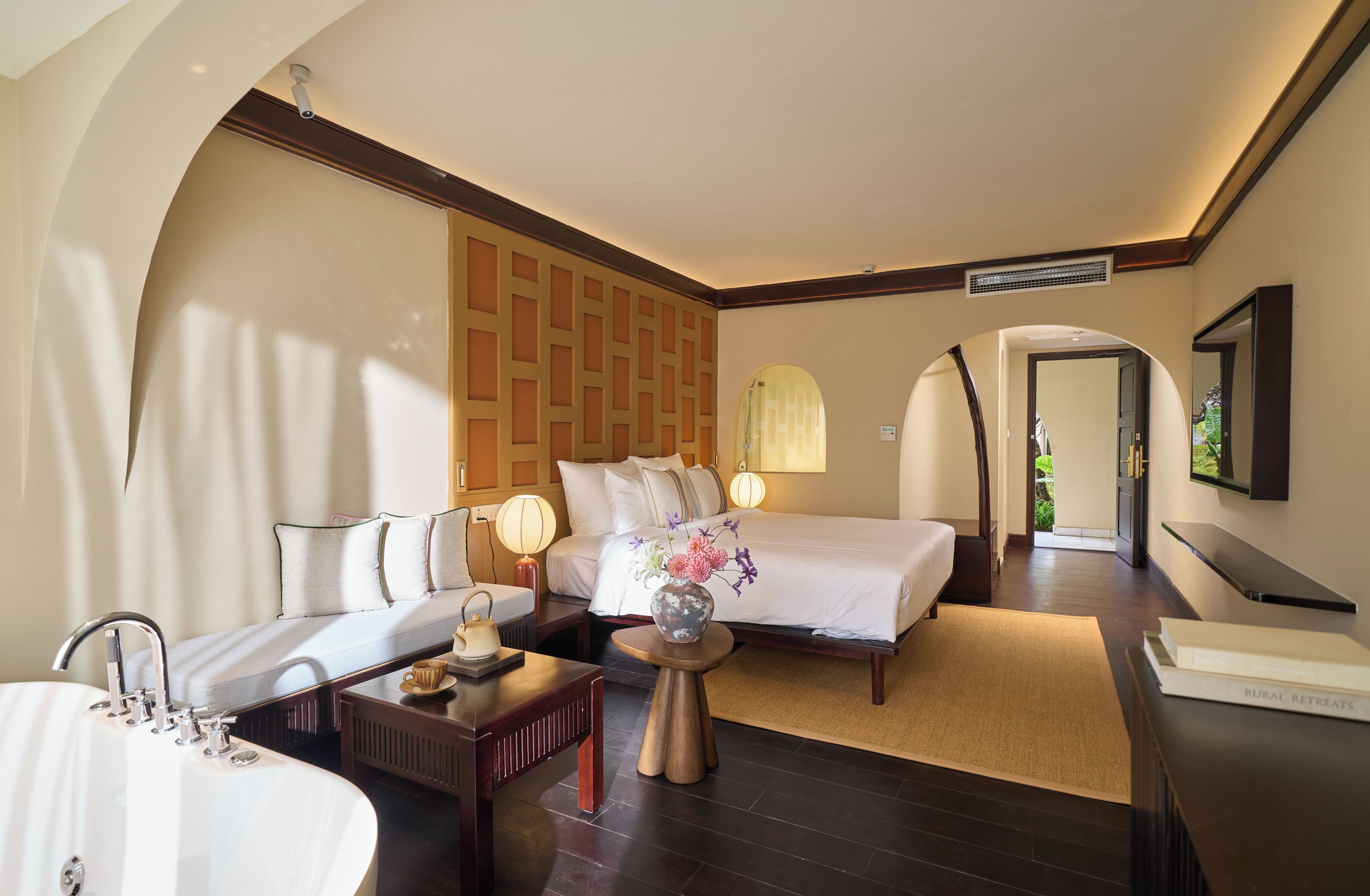 A hotel room with a bed, a sofa, a desk, a lamp, a vase with flowers, and a bathroom with a sink and mirror.