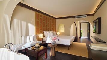 A hotel room with a bed, a sofa, a desk, a lamp, a vase with flowers, and a bathroom with a sink and mirror.