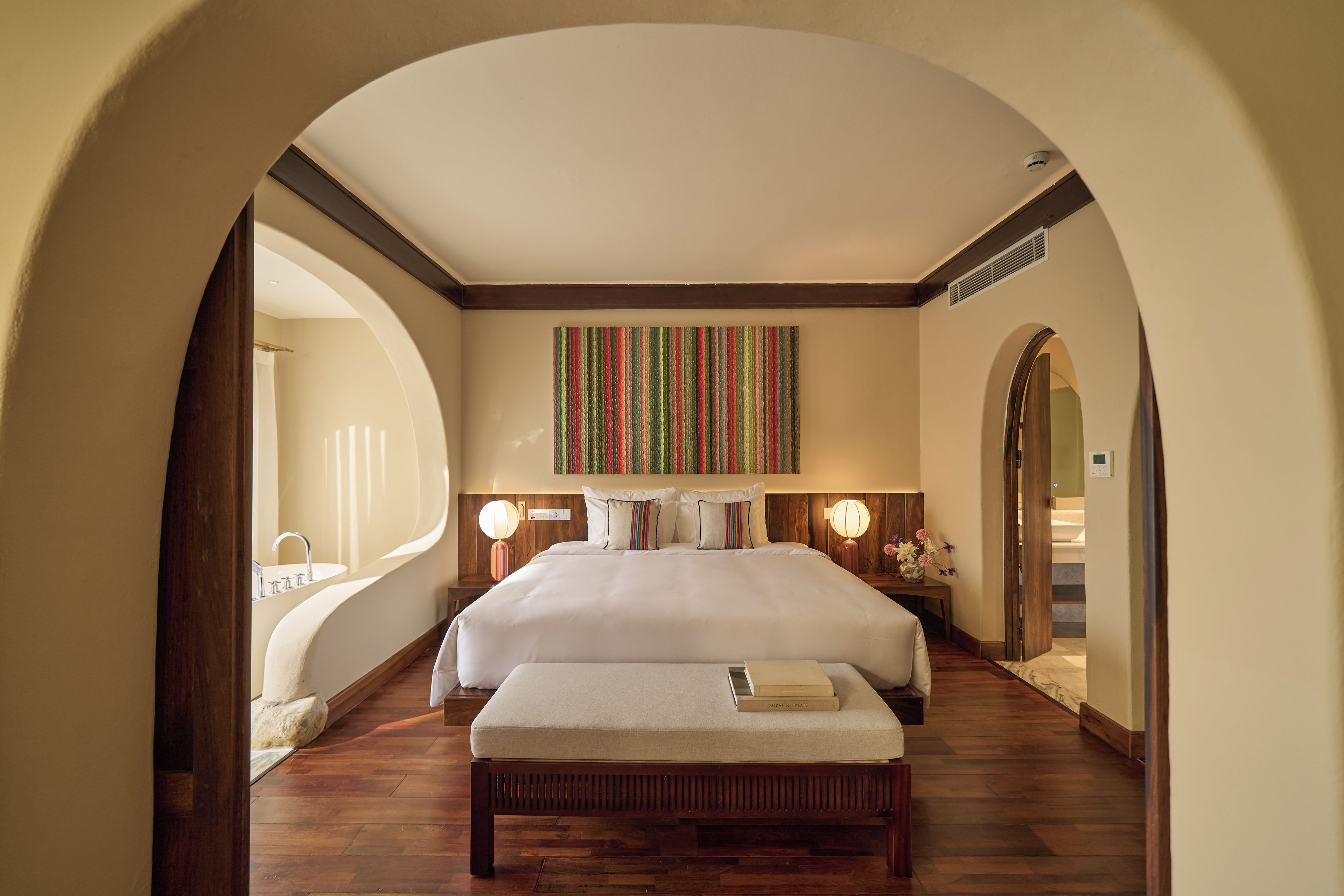 Habitación de hotel con una cama grande, un banco, una mesita de noche con lámpara y vistas a través de un arco.
