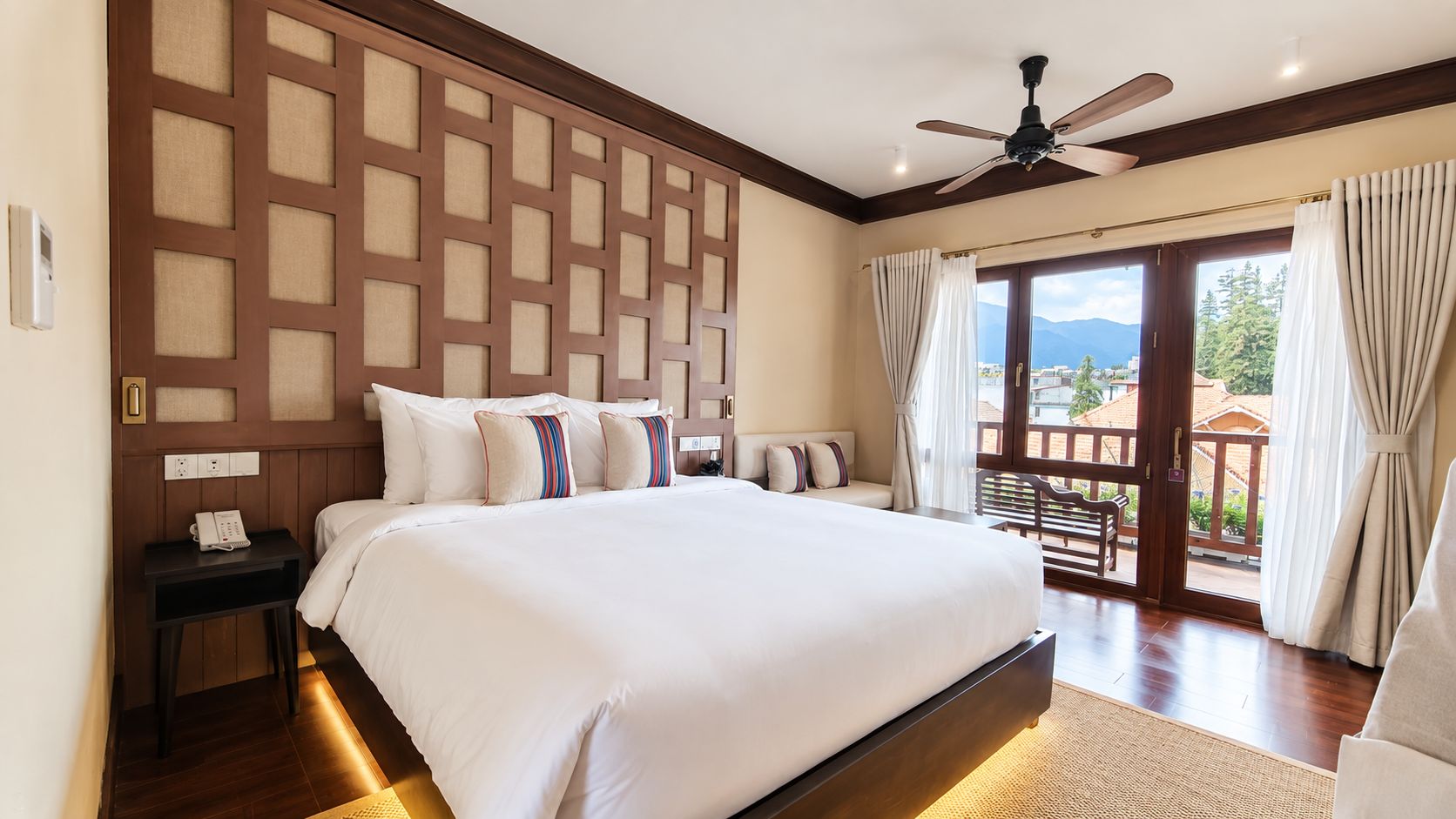 Habitación de hotel con una cama grande, cabecera de madera, ventilador de techo, mesita de noche con teléfono y vista al exterior a través de una puerta corrediza de vidrio.
