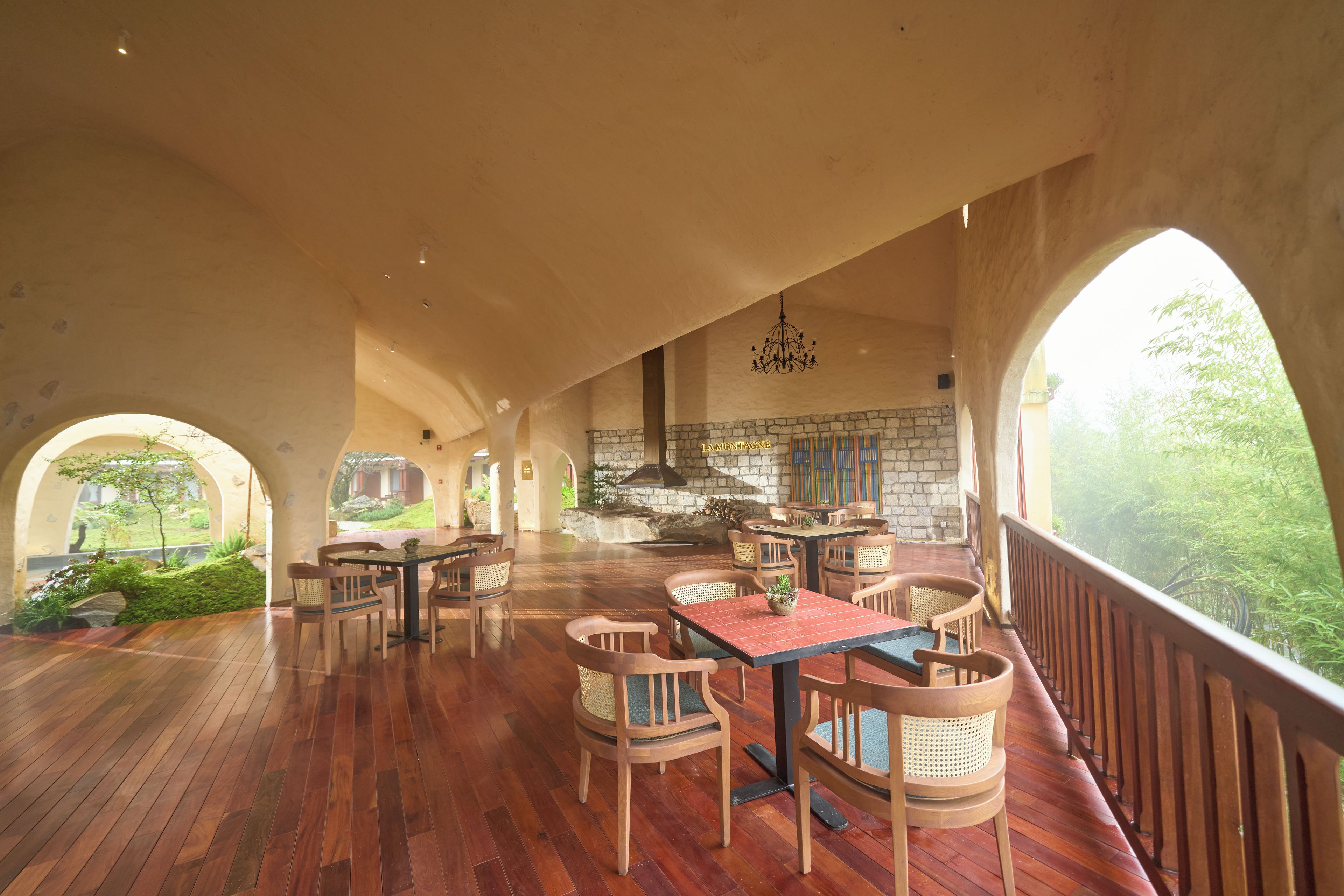 A spacious dining area with wooden flooring, multiple tables and chairs, a stone fireplace, and large arched windows offering a view of greenery.
