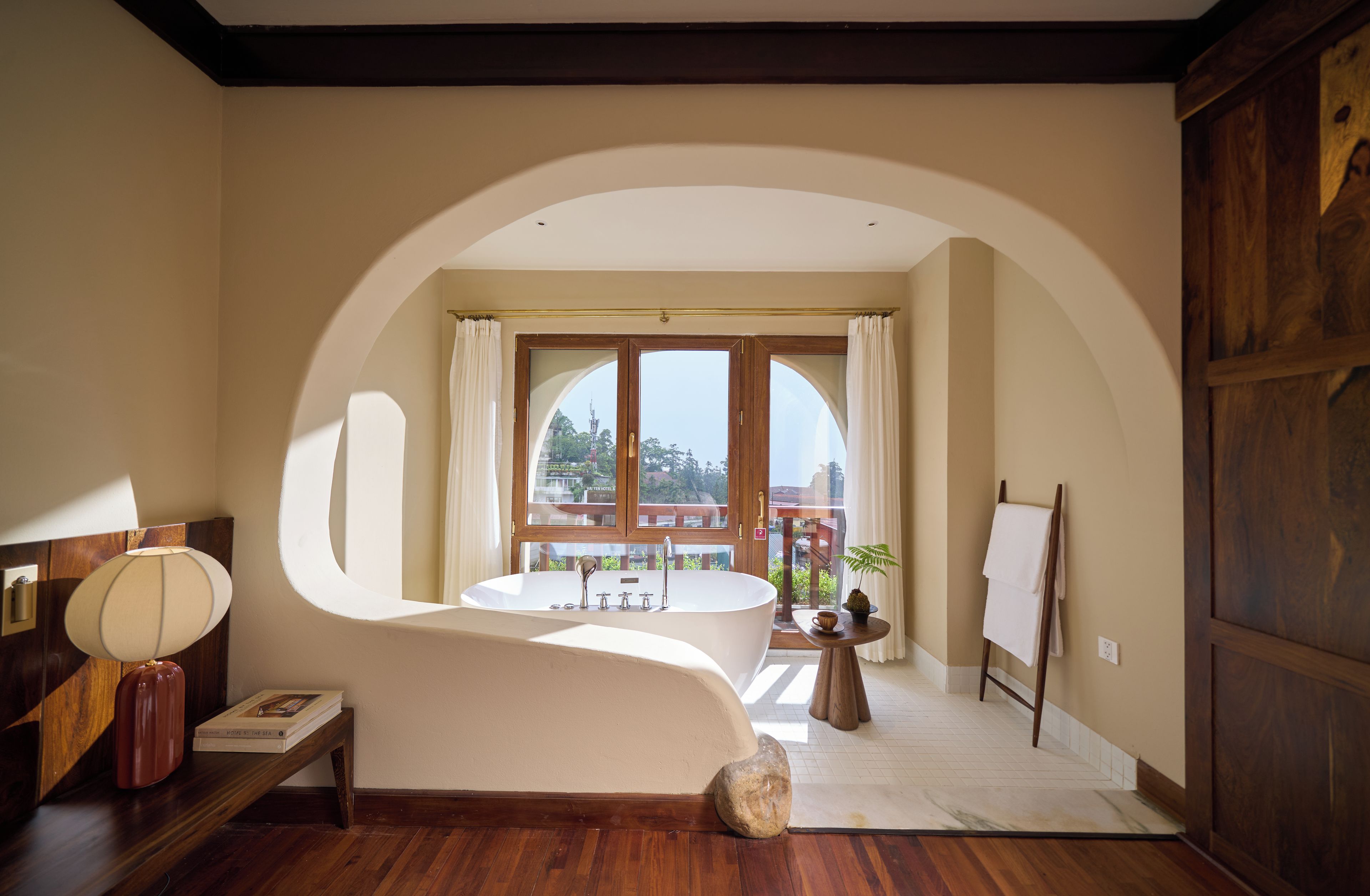 Un baño con una bañera independiente, un tocador de madera, un espejo redondo y vistas a un balcón con barandilla.