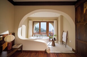A bathroom with a freestanding bathtub, a wooden vanity, a round mirror, and a view of a balcony with a railing.
