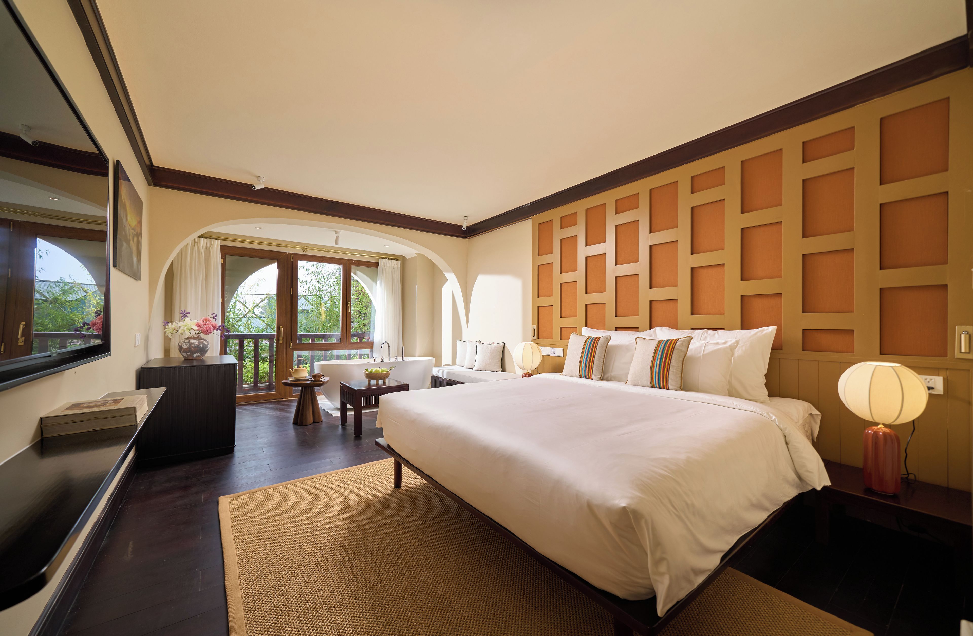 A hotel room with a large bed, a desk, a chair, a TV, and a view of greenery outside.
