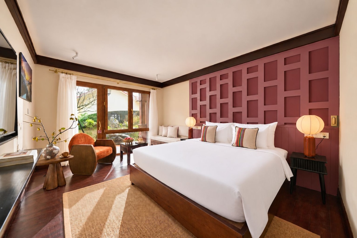 A hotel room with a large bed, a sitting area with a chair and table, a window with curtains, and a decorative wall panel.