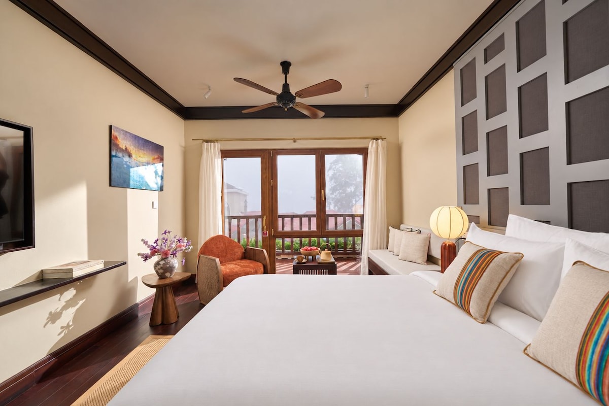 A hotel room with a large bed, a ceiling fan, a TV, a painting, a window with curtains, and a balcony with a view.