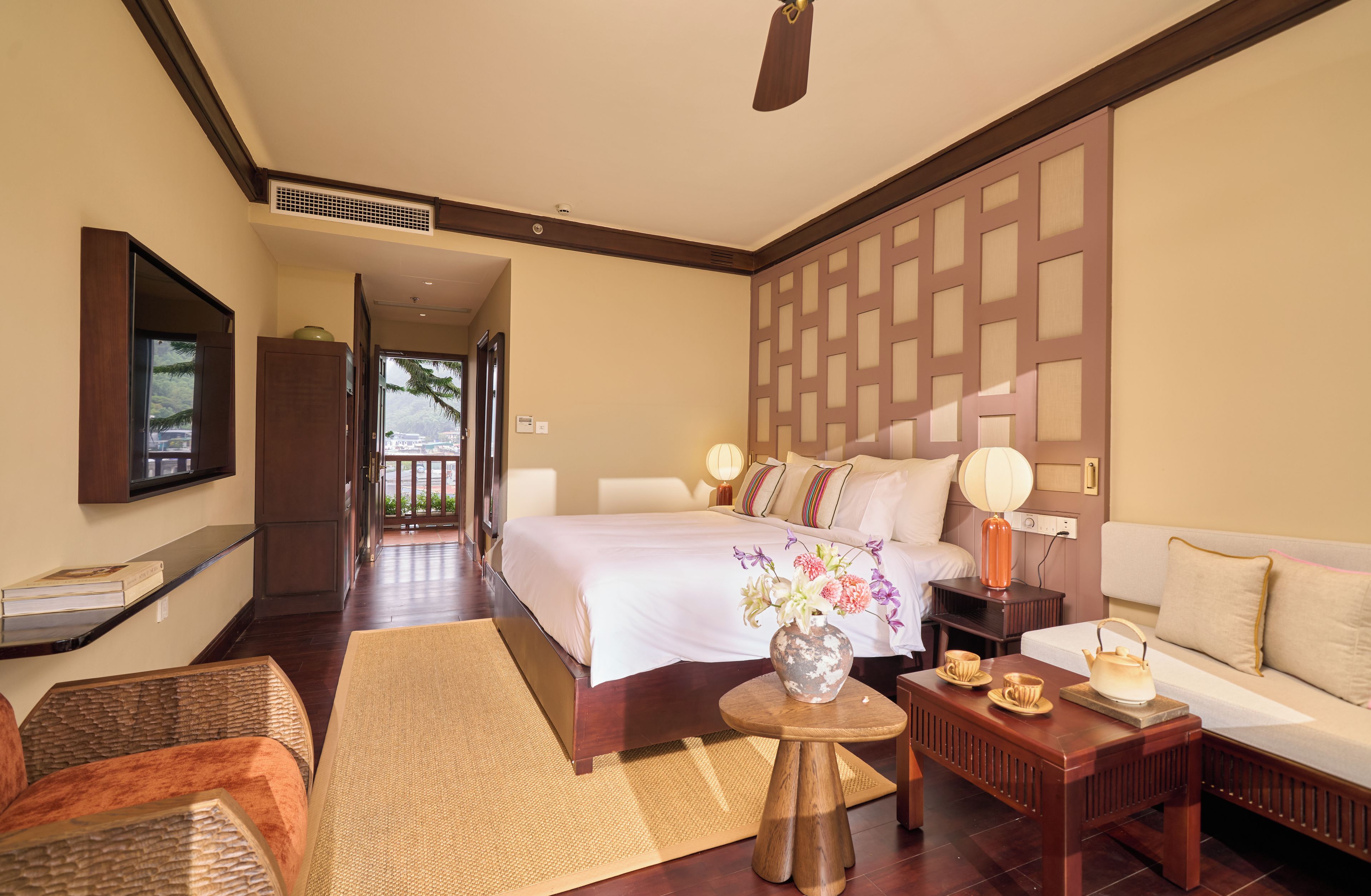 Habitación de hotel con una cama grande, un sofá, una mesita con un jarrón de flores y vistas al exterior.