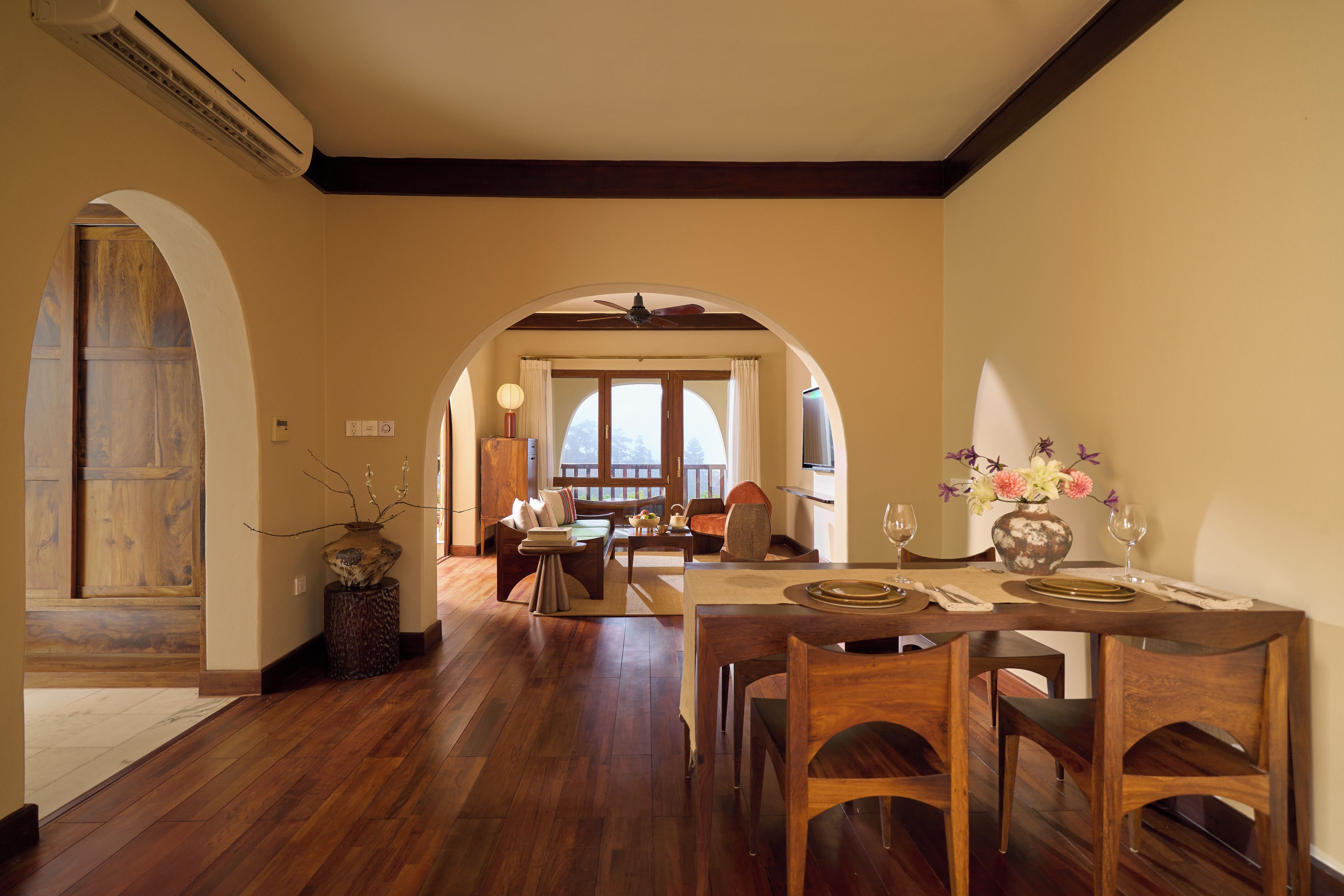 Un comedor con una mesa de madera dispuesta para dos, un jarrón con flores y vistas a un balcón por un arco.