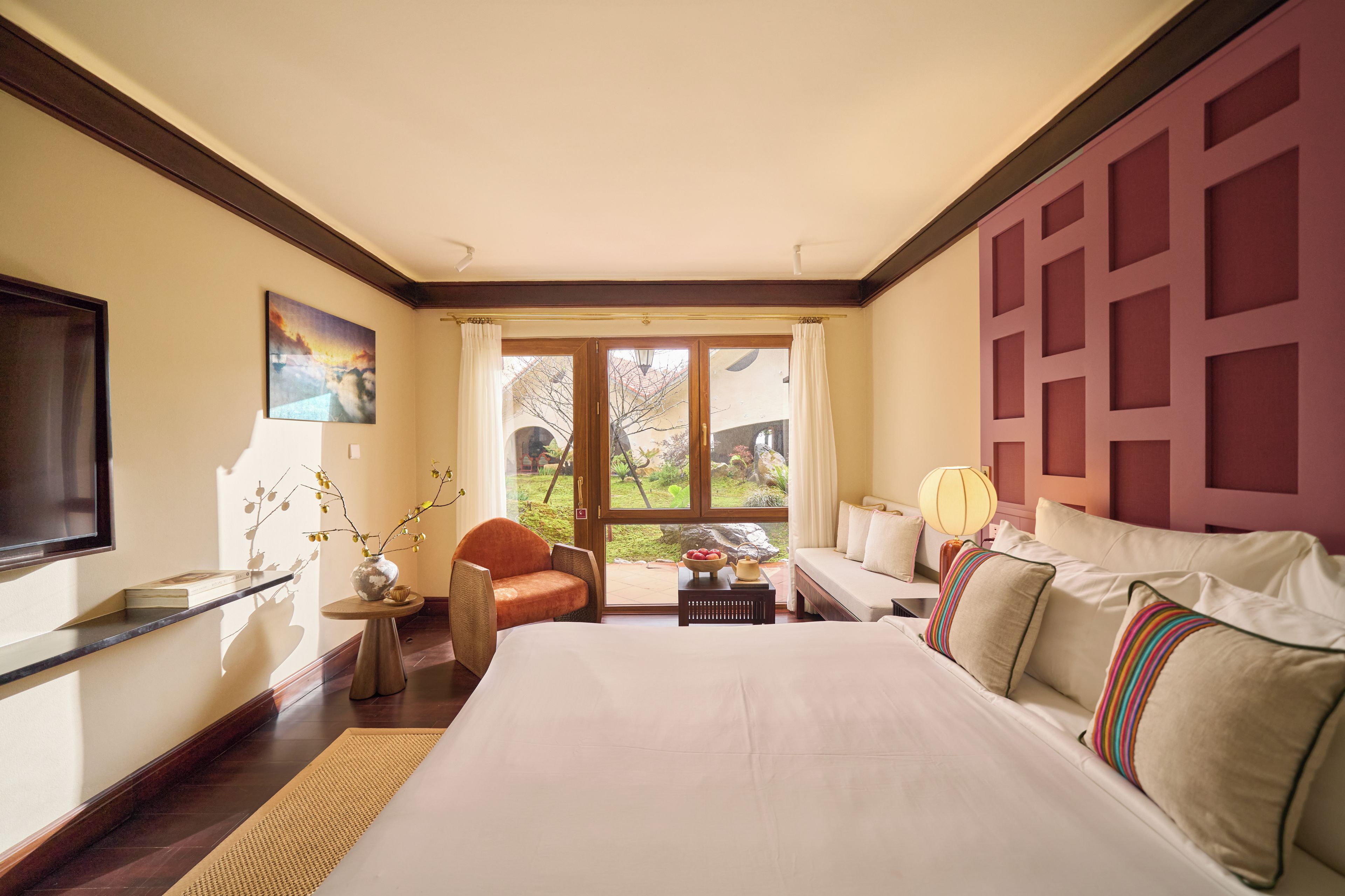 A hotel room with a bed, a chair, a table with a bowl of fruit, a vase with branches, a painting, and a view of the outdoors through a large window.