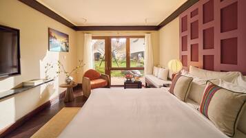 A hotel room with a bed, a chair, a table with a bowl of fruit, a vase with branches, a painting, and a view of the outdoors through a large window.