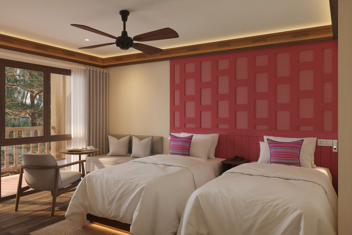 A hotel room with two beds, a ceiling fan, a small table with a chair, and a view of trees through a window.