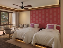 A hotel room with two beds, a ceiling fan, a small table with a chair, and a view of trees through a window.