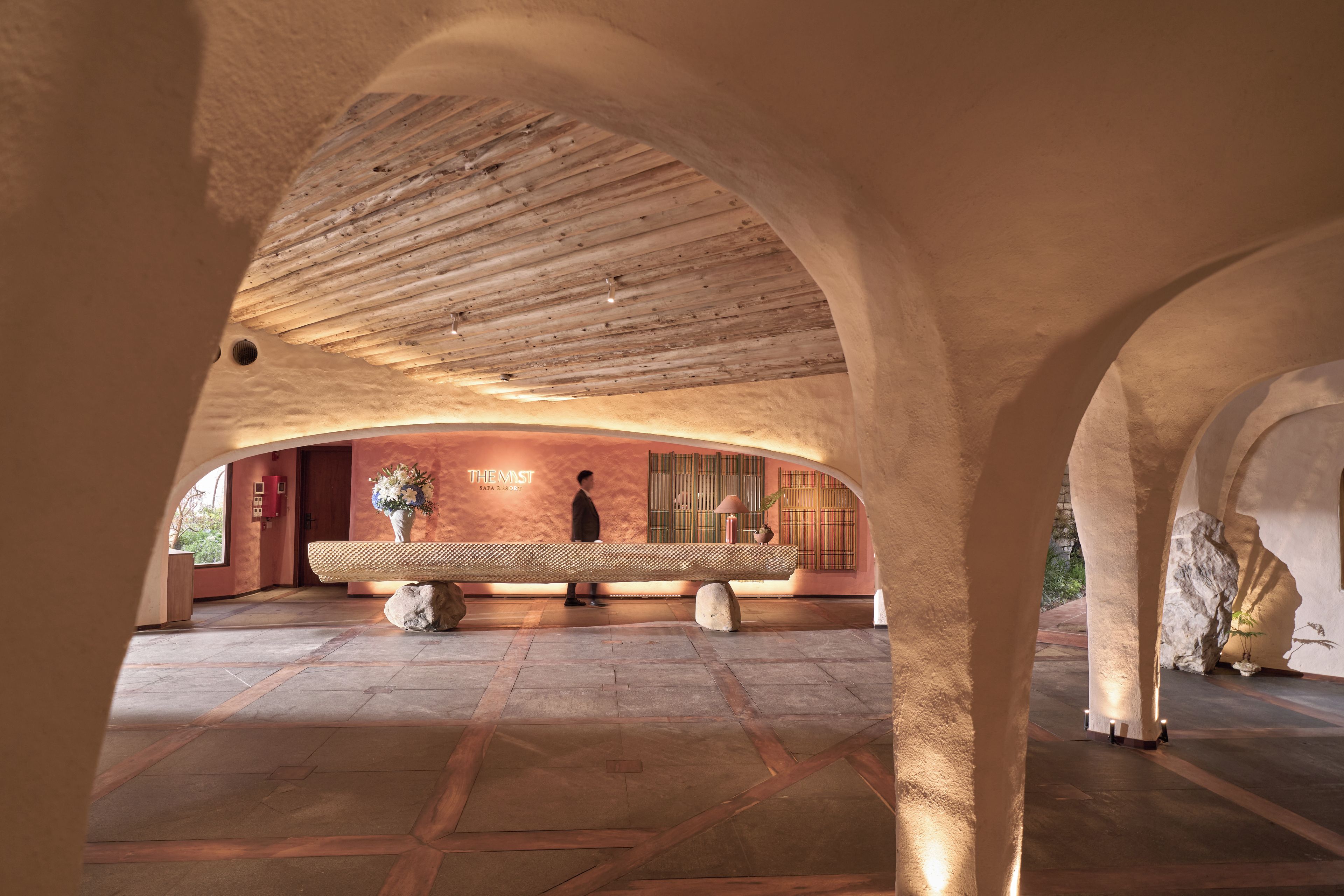 Un espacio interior con techo de madera, un banco de piedra y una pared con un arreglo floral.