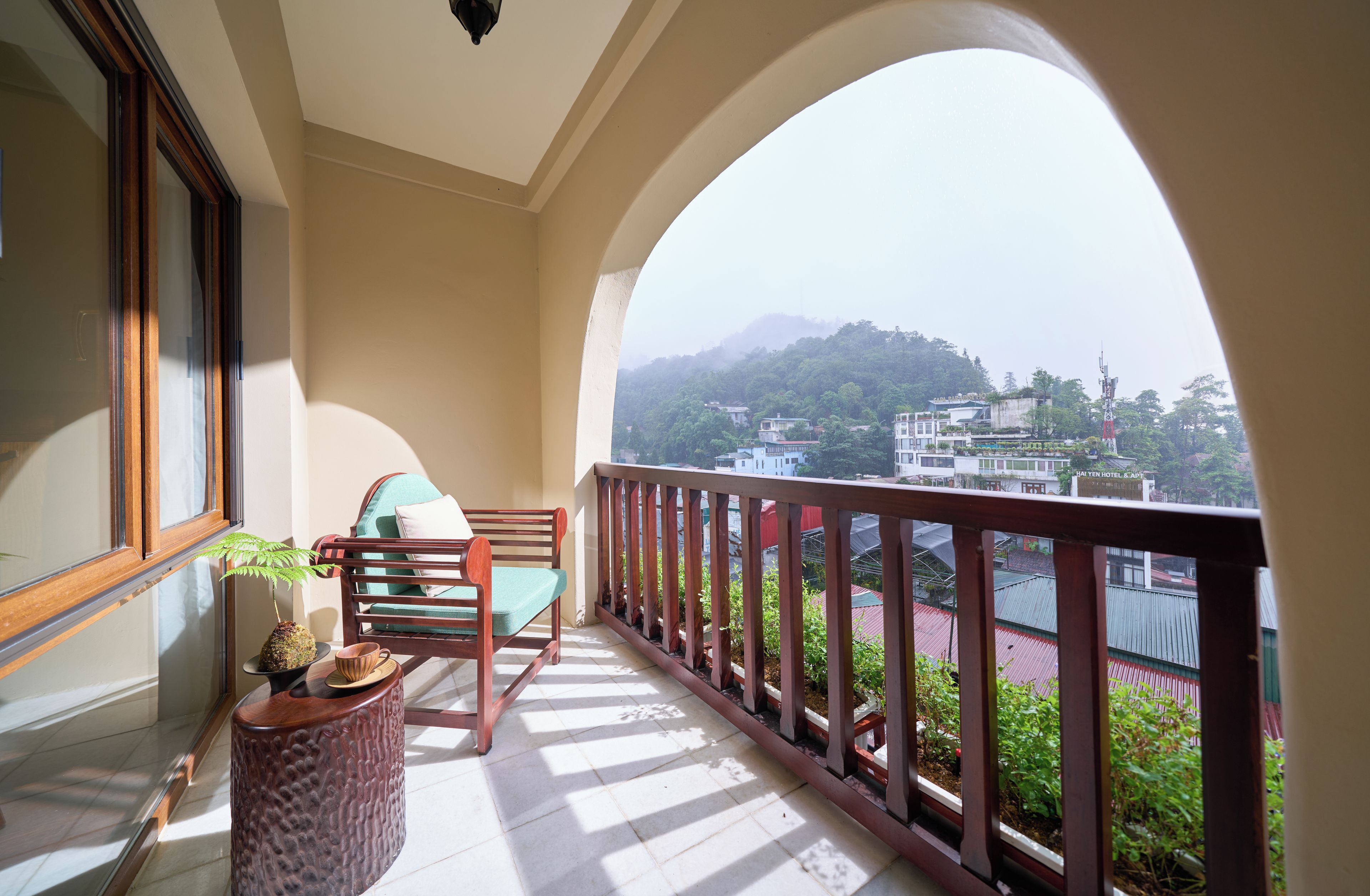 A balcony with a wooden chair, a small table with a plant, and a view of a hillside with buildings.