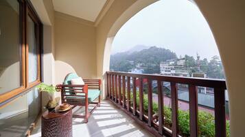 A balcony with a wooden chair, a small table with a plant, and a view of a hillside with buildings.