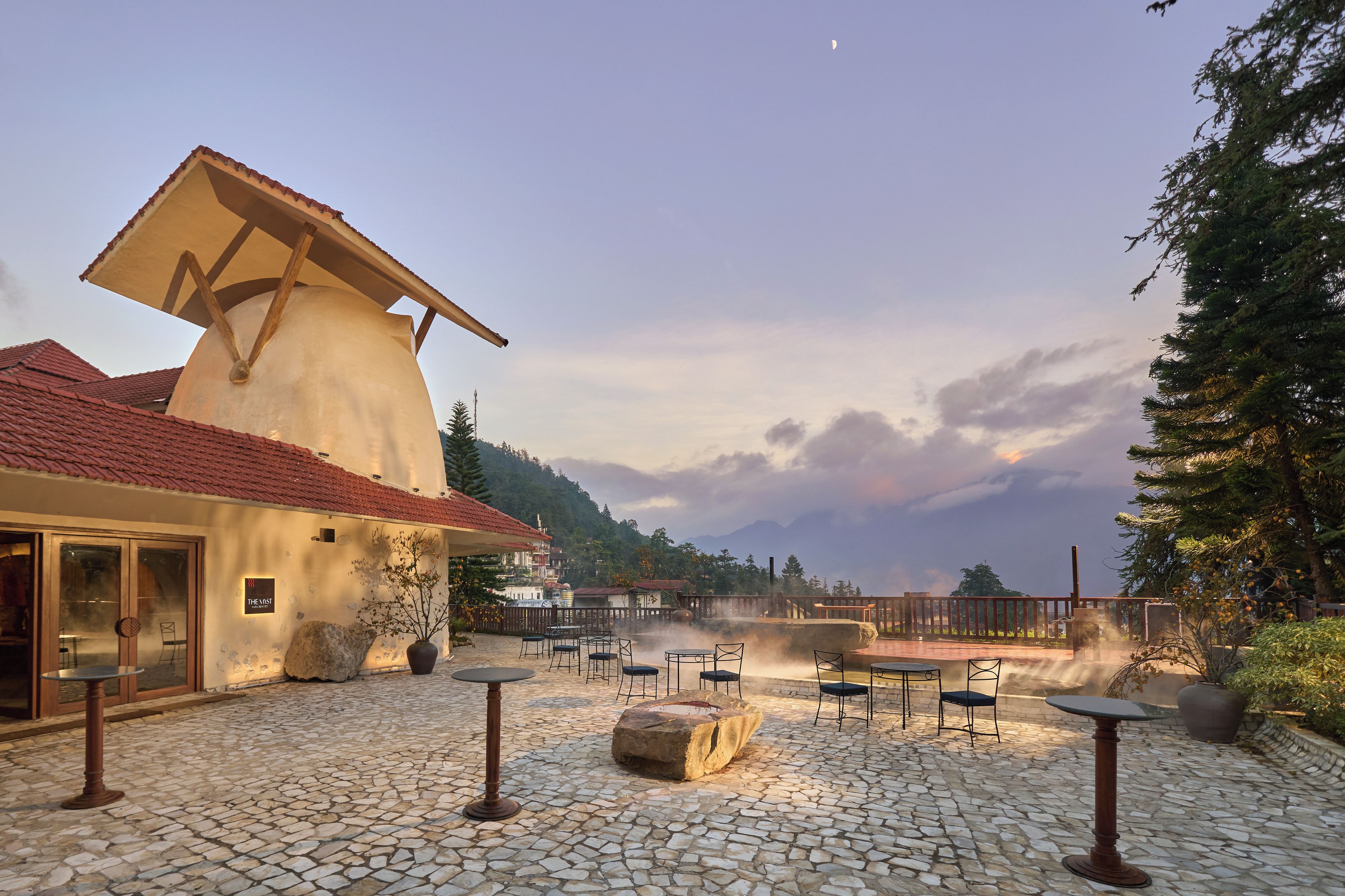 A courtyard with stone flooring, outdoor seating, and a building with a sloped roof.