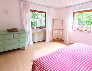 Ein Zimmer mit einem Bett, einem Schminktisch, einem Heizkörper, einem Wäscheständer und einem Fenster mit Vorhängen.