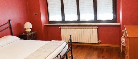 Une chambre à coucher comprenant un lit, une table de chevet, une commode, un radiateur et une fenêtre avec des stores.