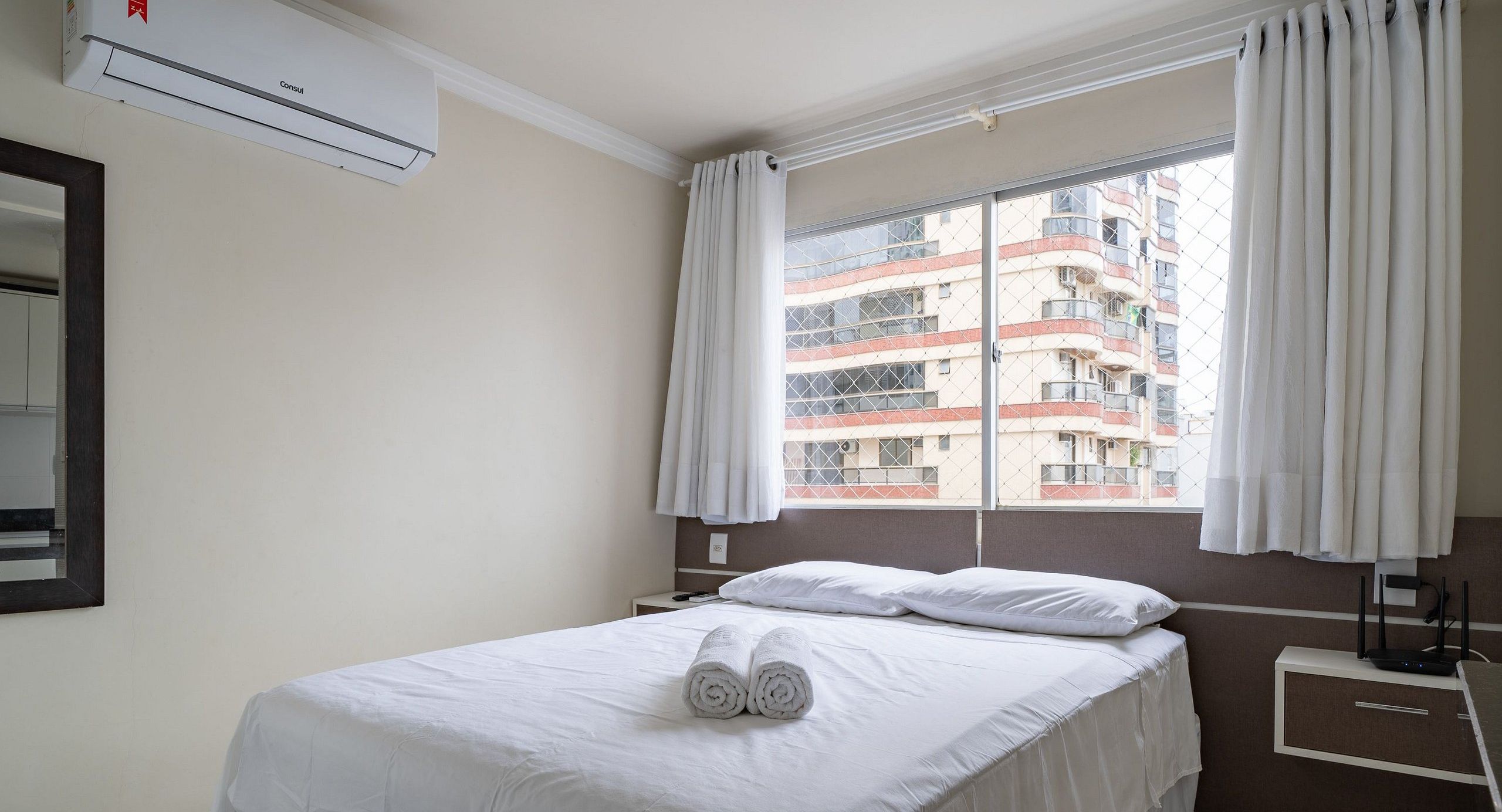 Une chambre d’hôtel comprenant un lit, une fenêtre avec des rideaux, un appareil de climatisation, une table de chevet et un miroir fixé au mur.