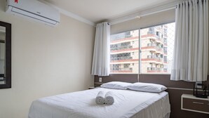 Habitación de hotel con cama, ventana con cortinas, equipo de aire acondicionado, mesita de noche y espejo montado en la pared.