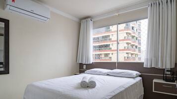Una camera d'albergo con un letto, una finestra con tende, un condizionatore d'aria, un comodino e uno specchio fissato alla parete.