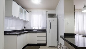 Una cocina con armarios blancos, encimera negra, refrigerador de dos puertas y una ventana con cortinas translúcidas.