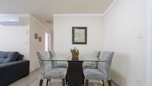 Un comedor con una mesa de vidrio, dos sillas estampadas, una obra de arte enmarcada en la pared y un sofá gris al fondo.