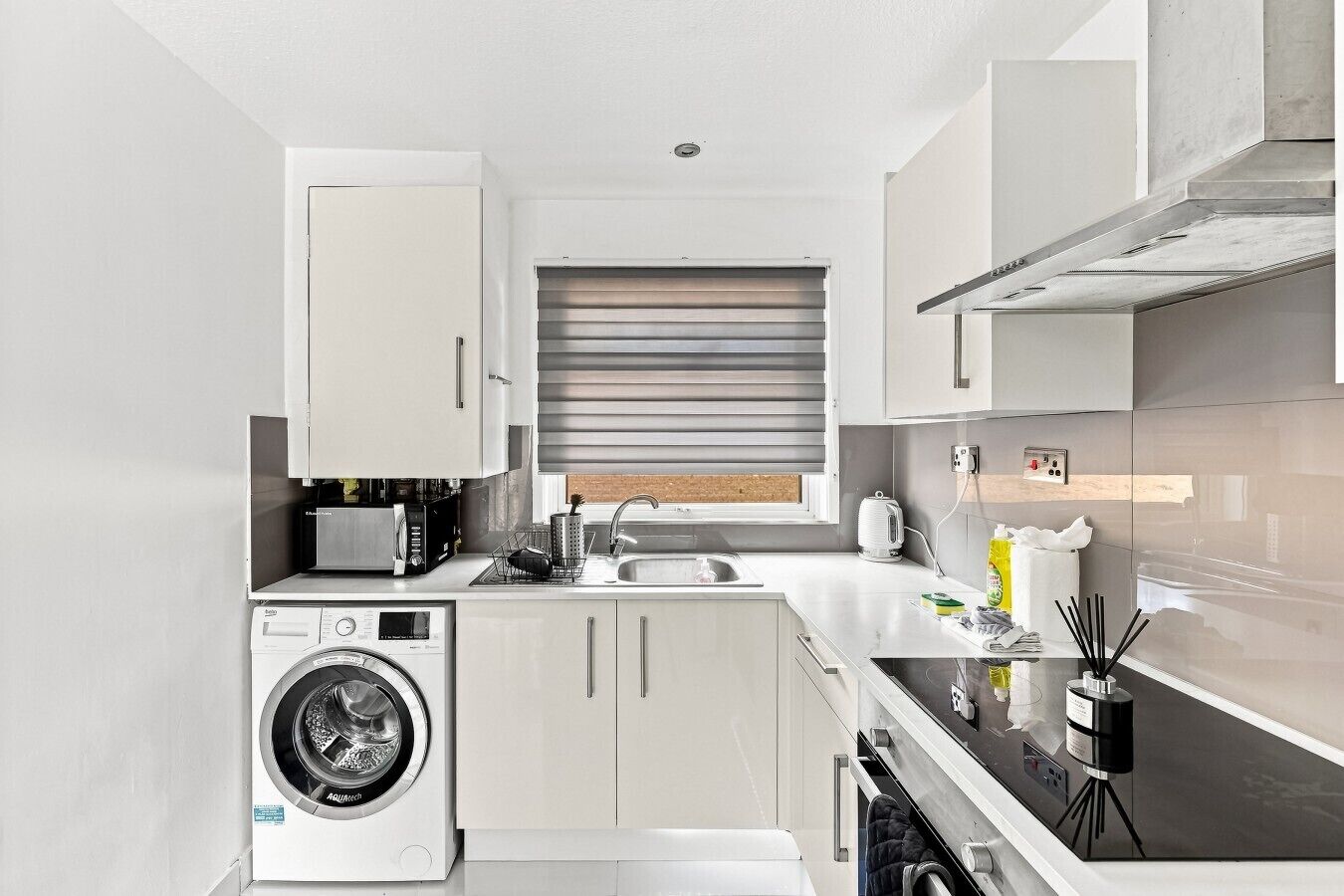 A modern kitchen with built-in appliances, a washing machine, and a window with blinds.
