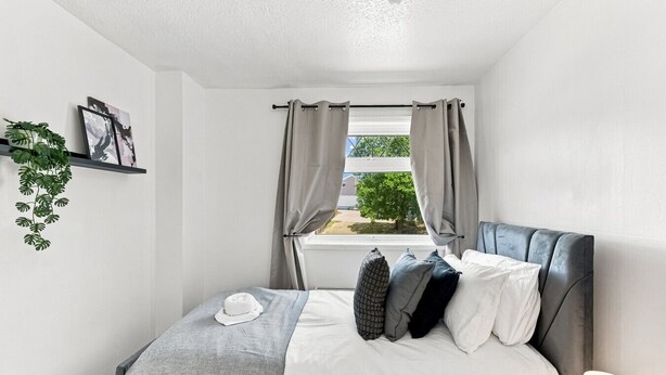 Ein Schlafzimmer mit einem Bett, einem Fenster mit Vorhängen, einer Pflanze an der Wand und einer Lampe auf dem Nachttisch.